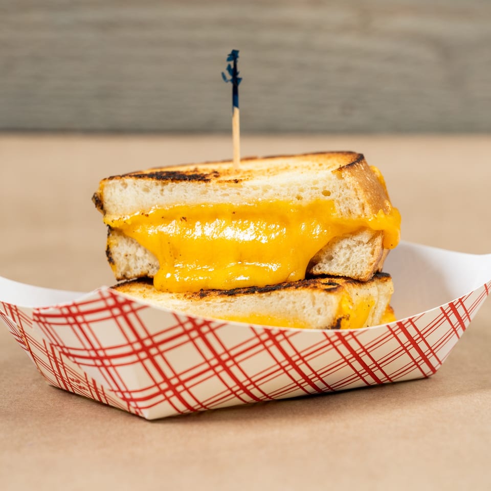 Grilled cheese sandwich with melted orange cheese in a red and white checkered paper tray.